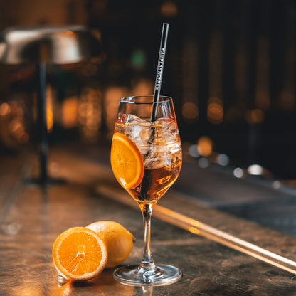 #IAMPLASTICFREE Trinkhalm 2.0 in Weißwein-Glas mit klarem, orangen Cocktail, Eiswürfeln und einer Orangenscheibe, dabei eine halbierte Orange