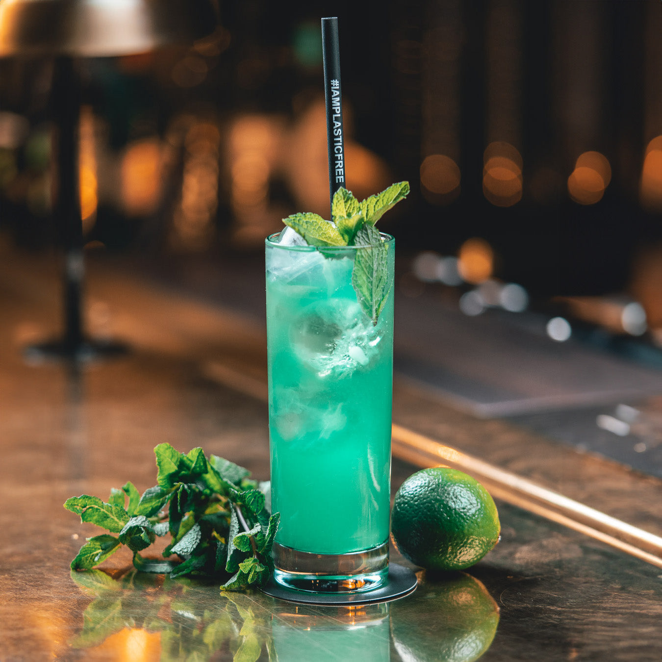 #IAMPLASTICFREE Trinkhalm in Highball-Cocktailglas mit türkisem Drink und Eiswürfeln, dabei Minze und Limette