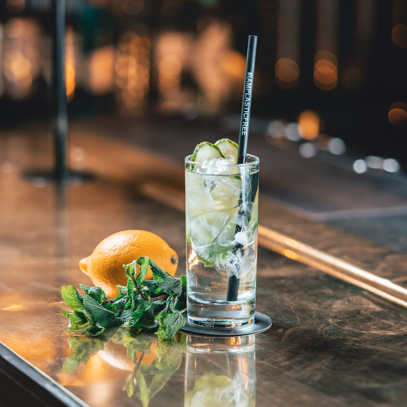 #IAMPLASTICFREE Trinkhalm 2.0 in Highball-Glas mit klarem Cocktail mit Eiswürfeln und Gurkenscheiben, dabei Minze und Orange