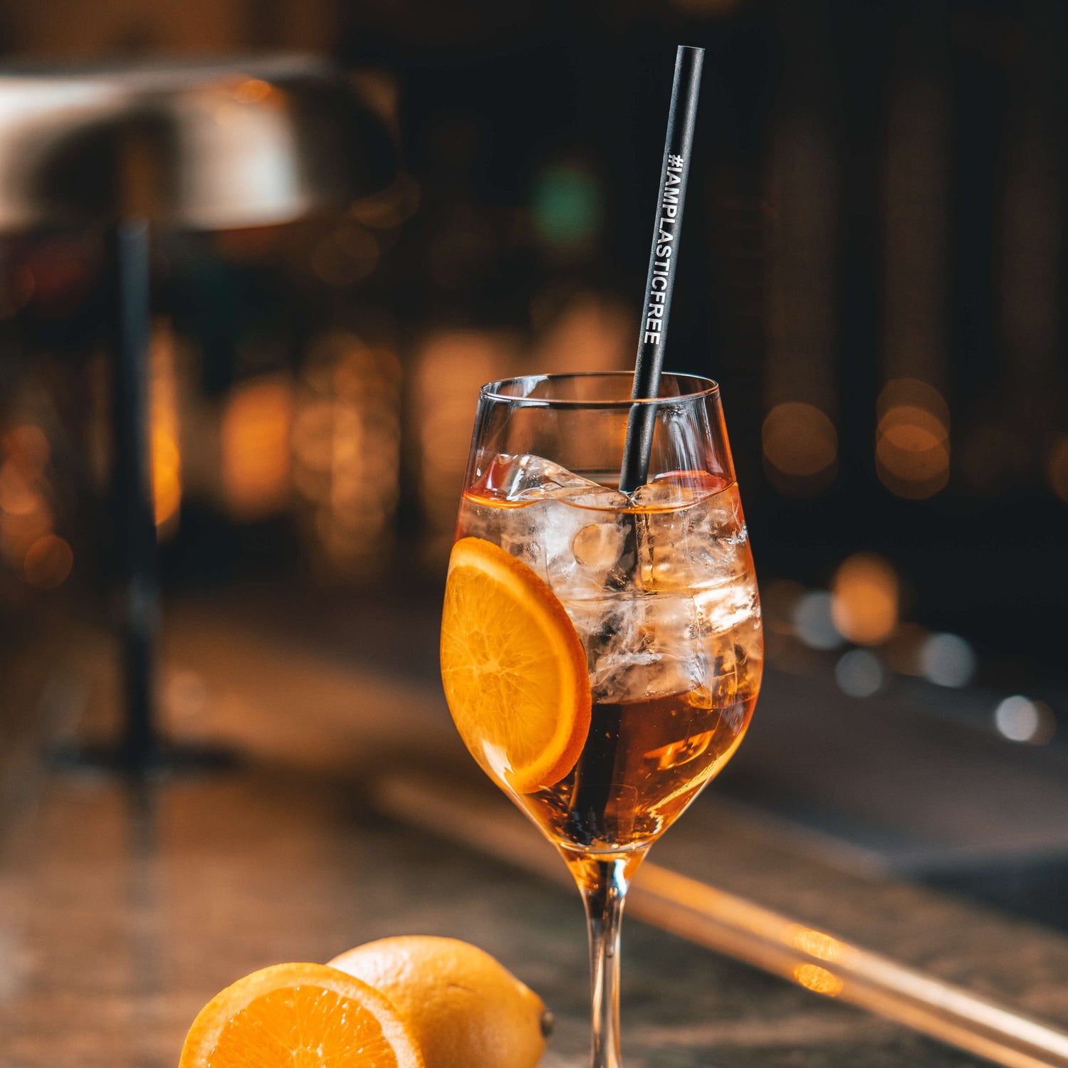 #IAMPLASTICFREE Trinkhalm 2.0 in Weißwein-Glas mit klarem, orangen Cocktail, Eiswürfeln und einer Orangenscheibe, dabei eine halbierte Orange