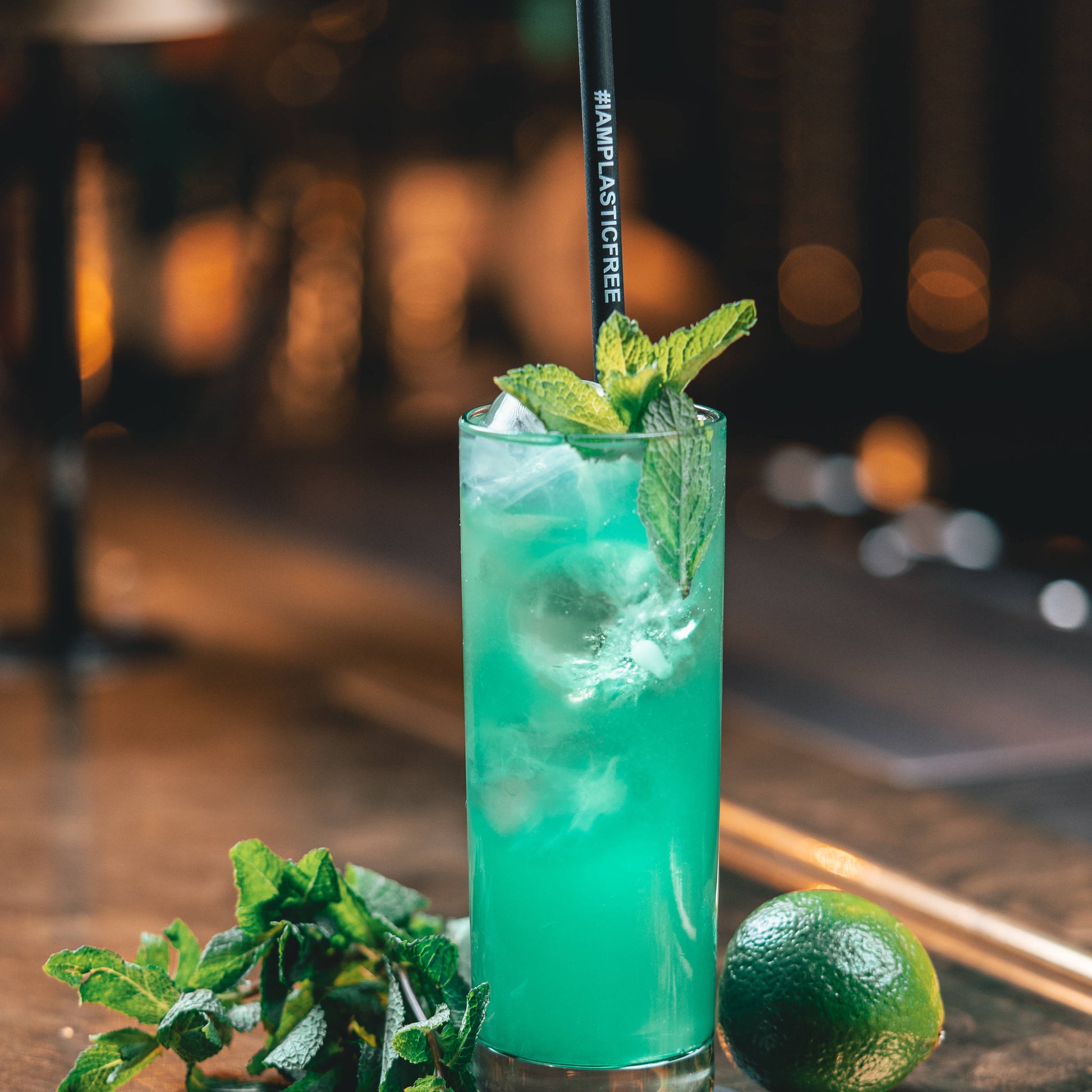 #IAMPLASTICFREE Trinkhalm 2.0 in Highball Glas mit türkisem Cocktail, Eiswürfeln und Minzblättern, dabei Limette und Minzzweig